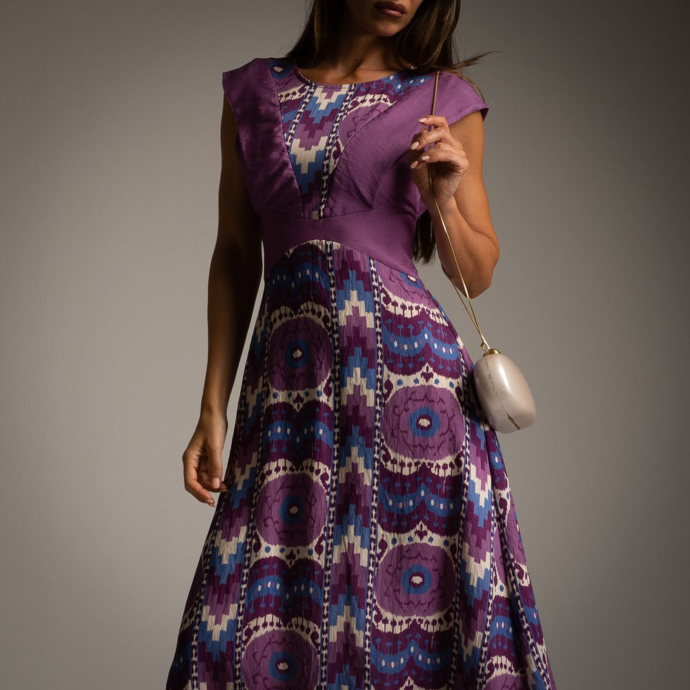 Close-up of purple midi dress with antique print and V-shaped bodice panels, styled with a clutch