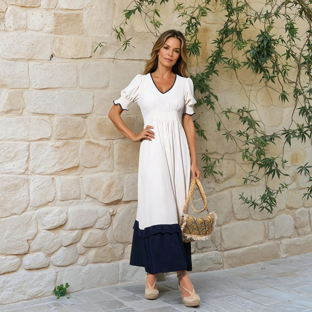 Elegant woman in off-white and navy midi dress standing by stone wall with greenery
