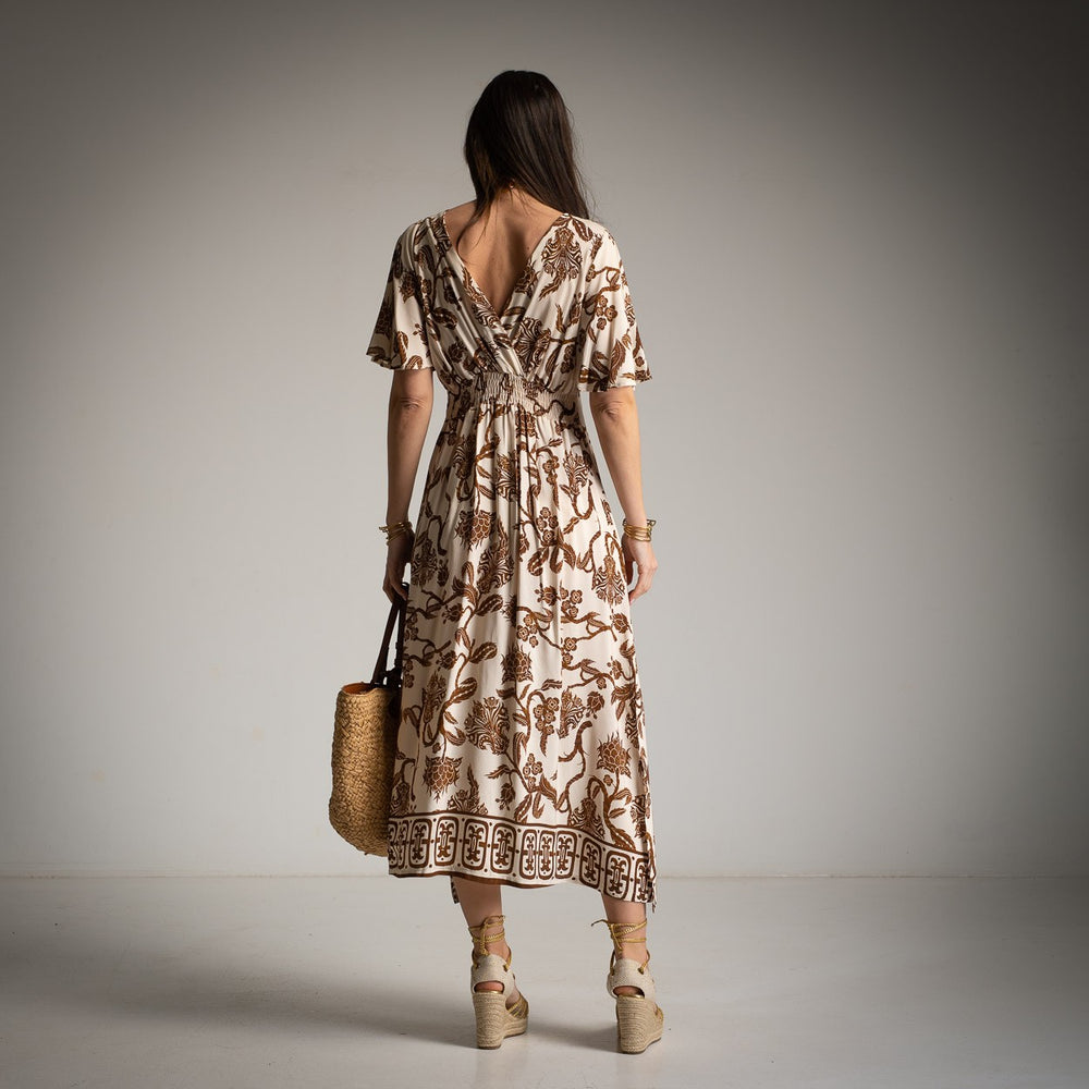 Back view of a woman wearing a beige and brown floral print dress with flutter sleeves, posing against a plain grey wall