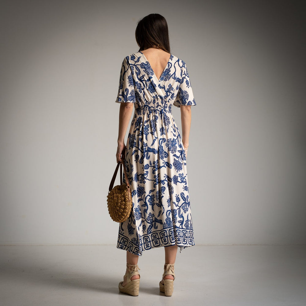 Back view of a woman wearing a beige midi dress with navy blue floral print and wide short sleeves, standing in front of a neutral grey background