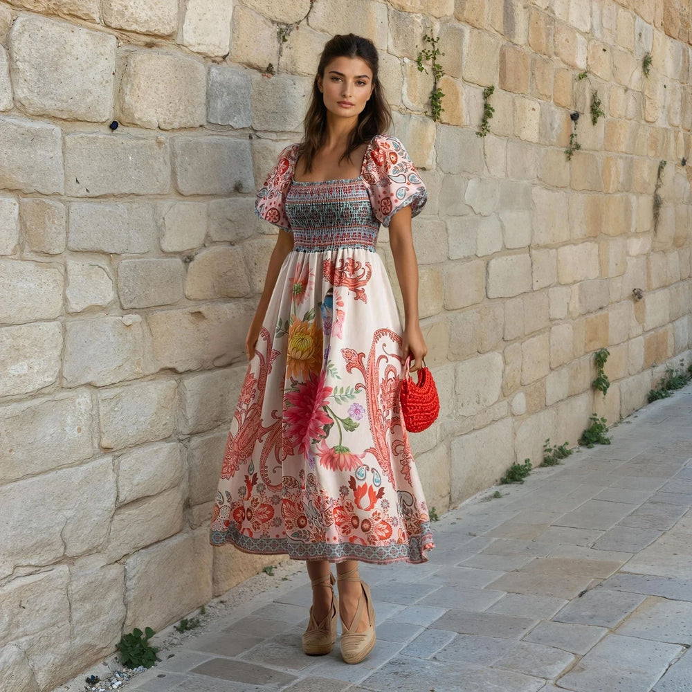 Model wearing a colorful floral-print midi dress with puff sleeves and a smocked bodice, styled with espadrilles and a red woven handbag, posing in front of a stone wall.