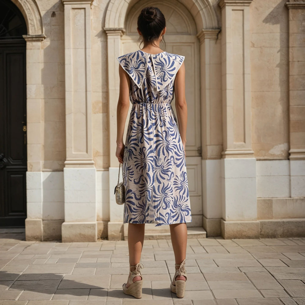 Back view of a sleeveless blue and beige print midi dress with gathered waist detail, shown outdoors in a city setting – MariGiou Elegant Daywear.