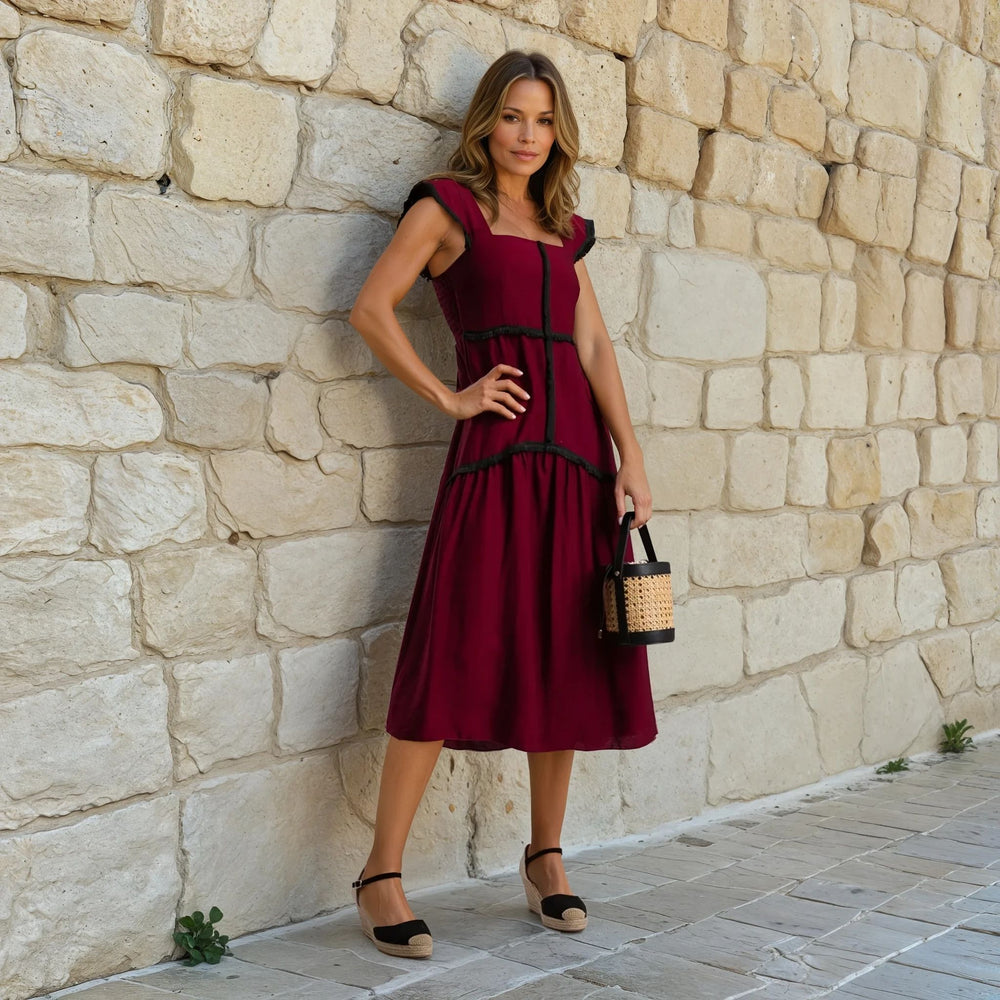 Woman posing in a sleeveless burgundy midi dress with decorative black fringe and square neckline, standing against a rustic stone wall.