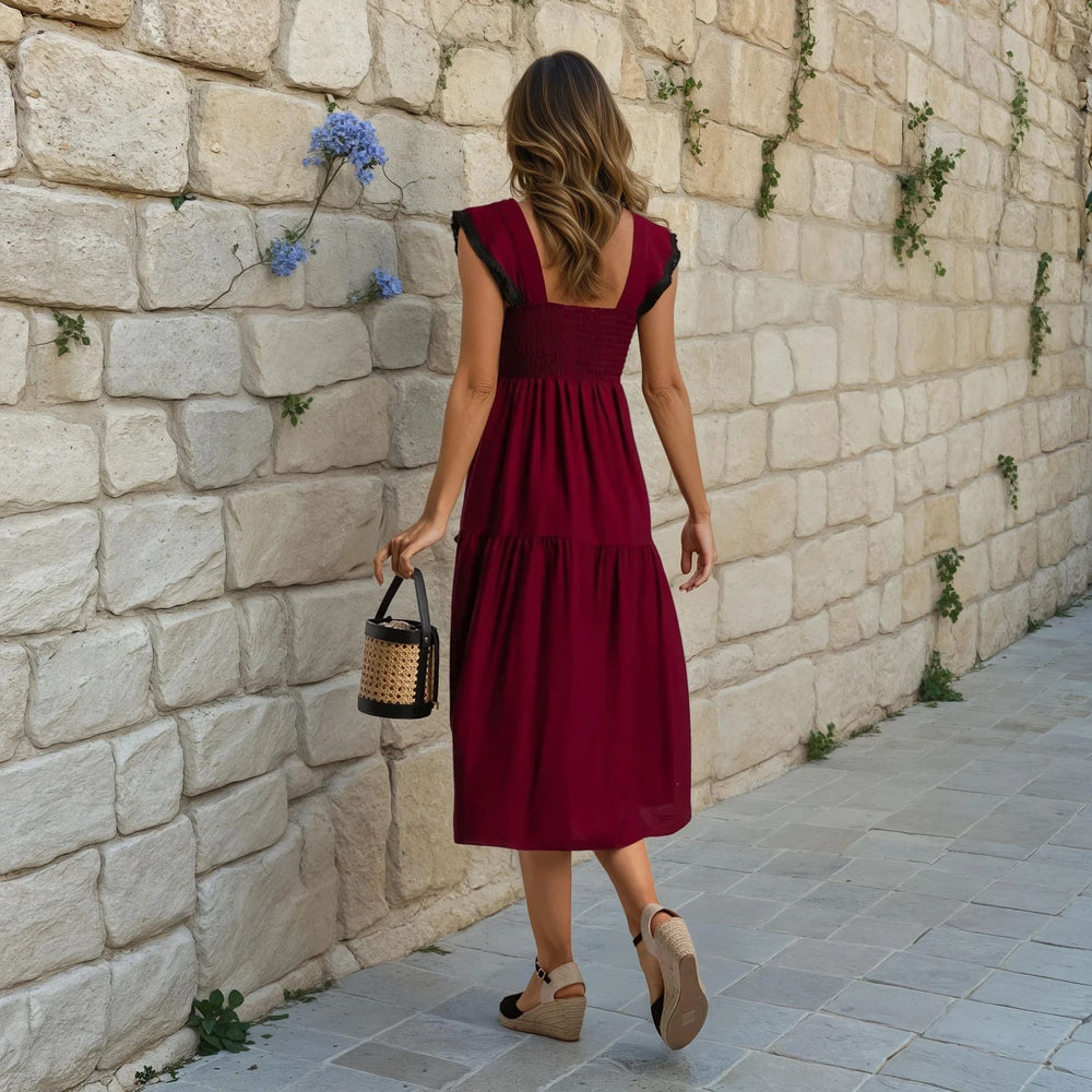 Back view of a woman wearing a sleeveless burgundy midi dress with black fringe trim, walking along a stone wall in summer wedges.