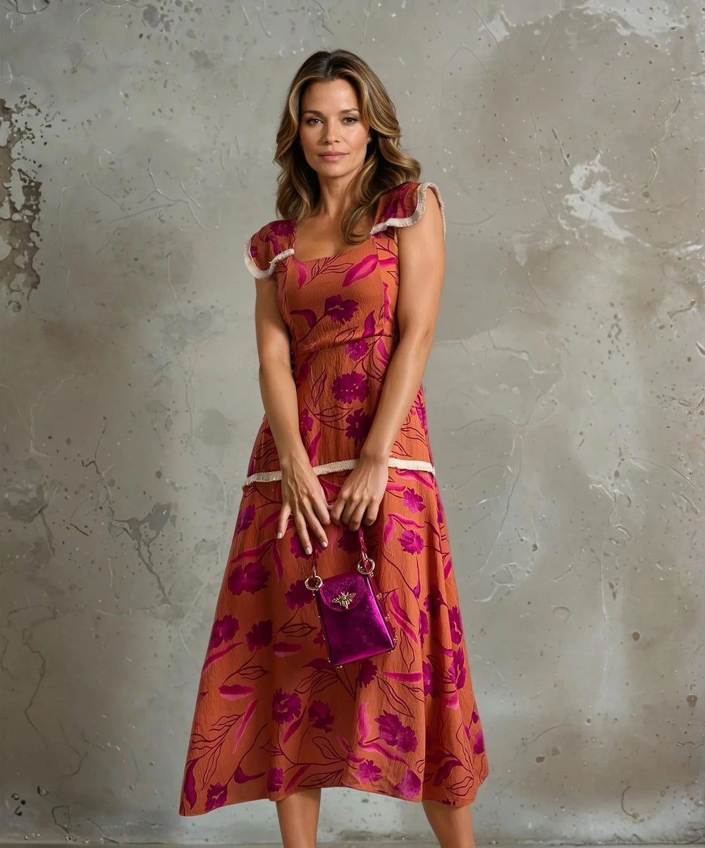 Close-up of a woman in a rust-orange dress with pink floral print and white-trimmed sleeves, holding a glossy pink handbag in front of a concrete background.