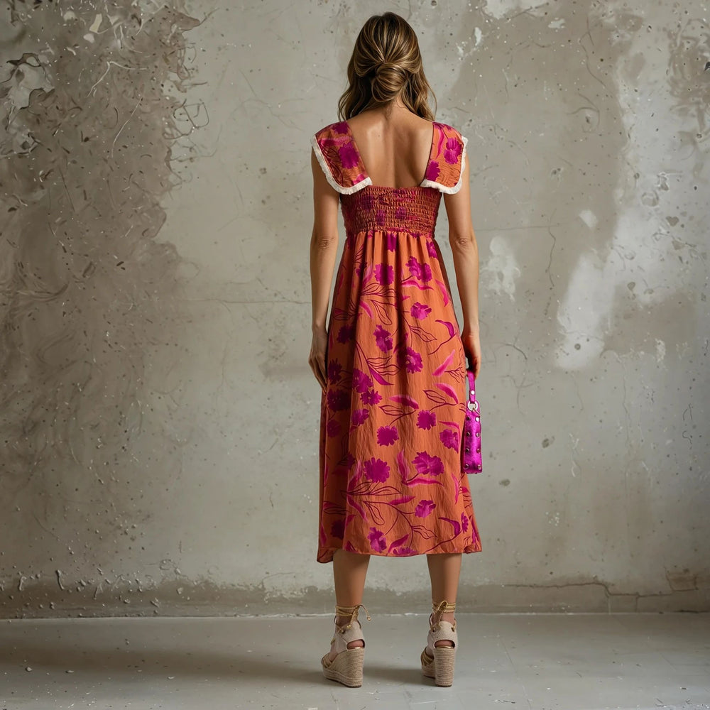 Back view of a woman in a rust-orange midi dress with pink floral design and smocked bodice, featuring white-trimmed cap sleeves, styled with wedge espadrilles.