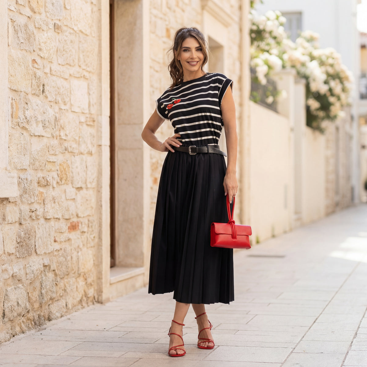 Model wearing black striped cherry print statement T-shirt by MARIΓIOÚ styled with a black pleated midi skirt, red heels and red clutch in a Mediterranean street setting.