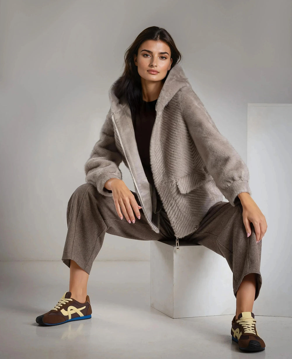 Model wearing MARIΓIOÚ faux fur hooded jacket in neutral tones, styled with tailored brown trousers.