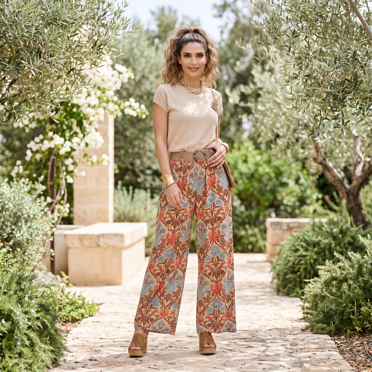 Woman wearing wide-leg pants with a baroque floral print and matching belt in a Mediterranean garden setting.